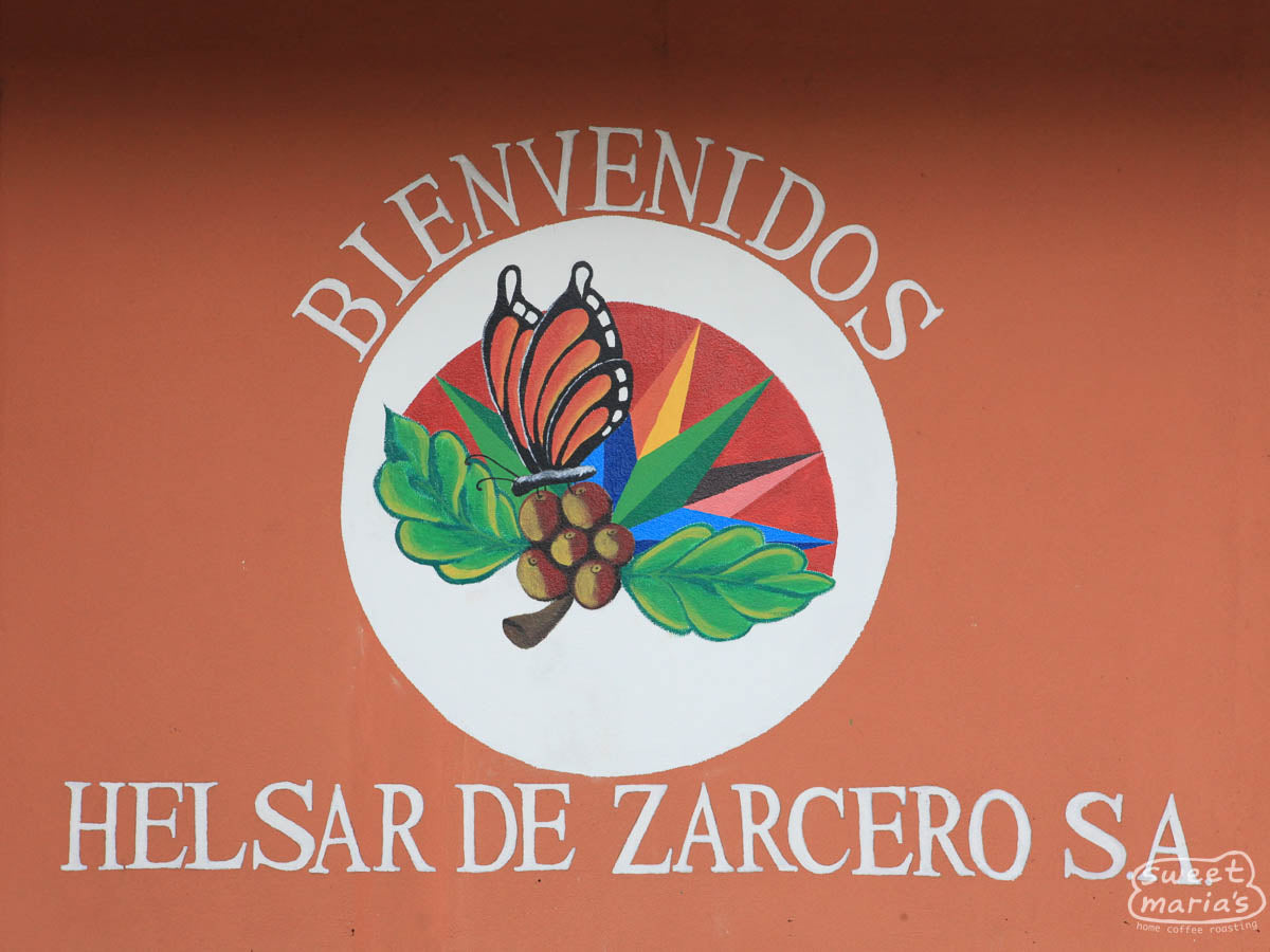 The colorful hand-painted signe reads, "Bienvenidos Helsar de Zarcero SA", with an image of a butterfly on a coffee tree.
